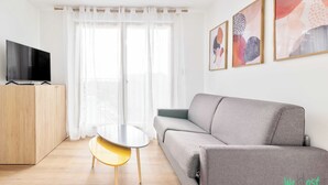 A modern living room with a grey sofa, a TV on a wooden cabinet, a small round table, and a window with sheer curtains.