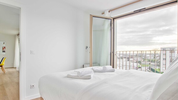 A bedroom with a large bed, a bathroom with a glass door, and a balcony with a view of the city.
