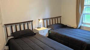 A small, simple room with two single beds, a nightstand with a lamp, and a window with curtains. - The Holy Family of Nazareth (Cullman)