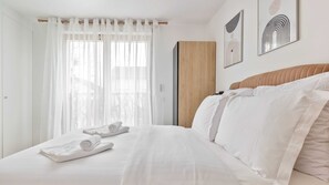Hotellihuone, jossa on sänky, valkoiset verhot, puinen vaatekaappi ja kaksi kehystettyä taideteosta seinällä.