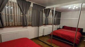 Une chambre avec un placard à portes miroir, deux lits avec une literie rouge, un radiateur et une fenêtre avec des rideaux gris.