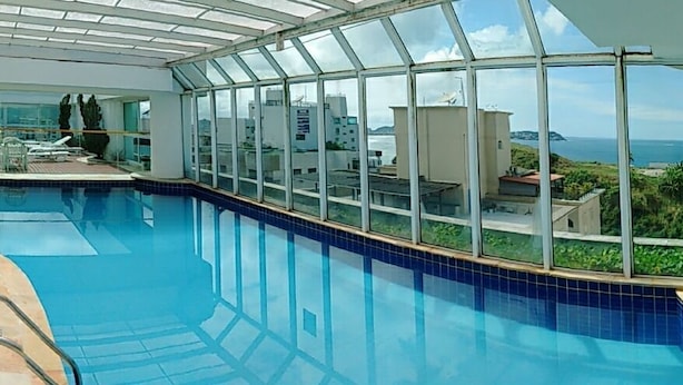 An indoor swimming pool with clear glass walls, a tiled floor, and a view of the sea and buildings. - Apê da Mi (Guaruja)