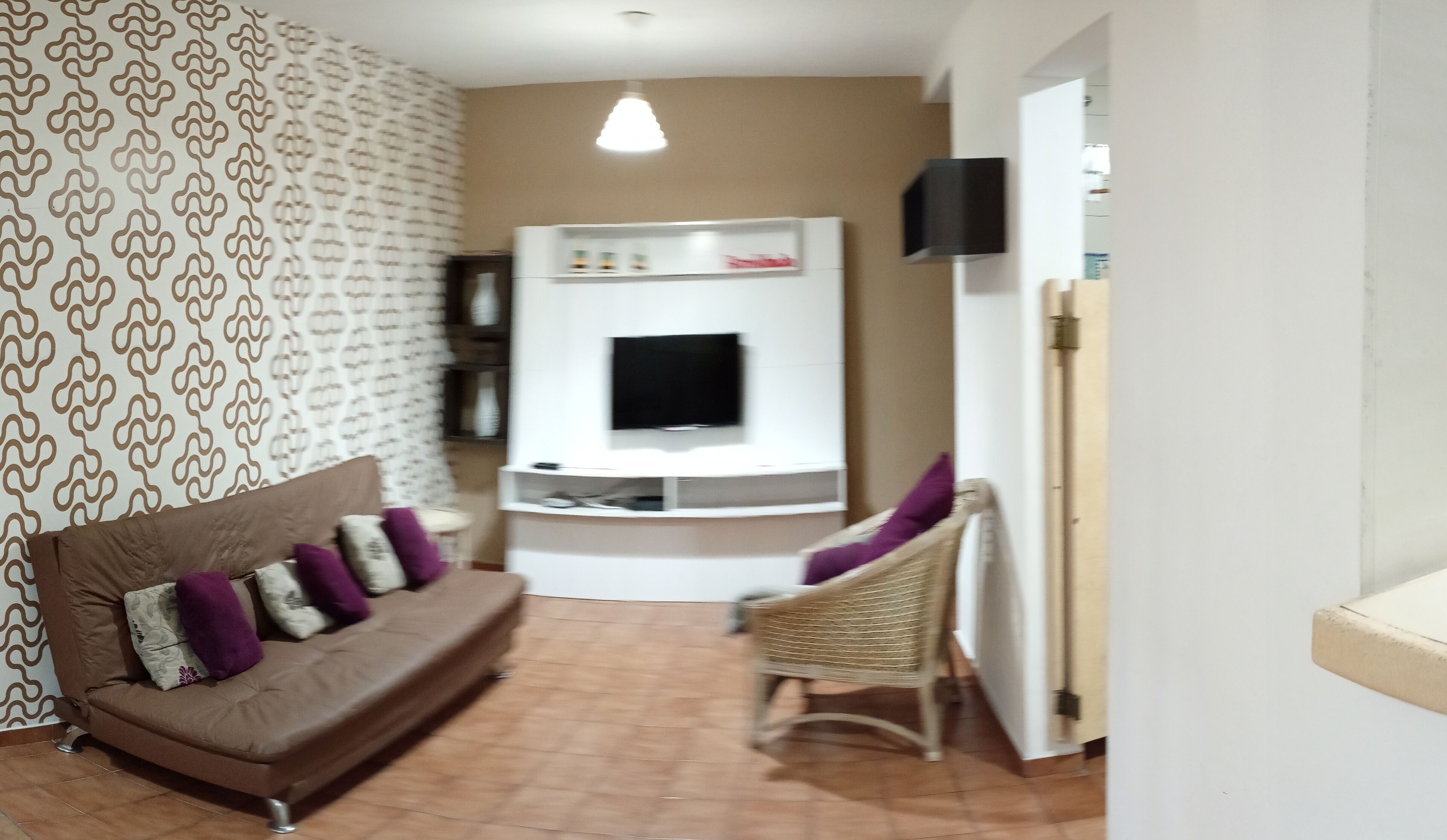 A living room with a brown sofa, a wicker chair, a flat-screen TV mounted on a white wall, and patterned wallpaper.