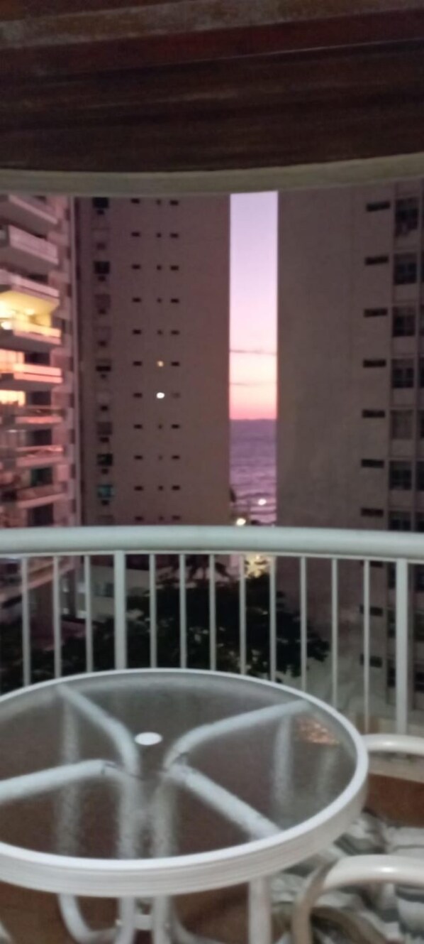 A balcony with a glass table and chairs, overlooking a cityscape with tall buildings and a sunset. - Apê da Mi (Guaruja)