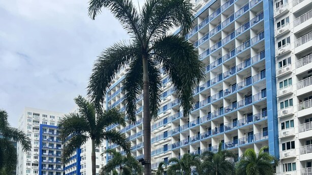 A resort with a swimming pool, palm trees, and a large building with balconies. - MOA-View 1BR - 5 min Walk to MOA Arena (Pasay)