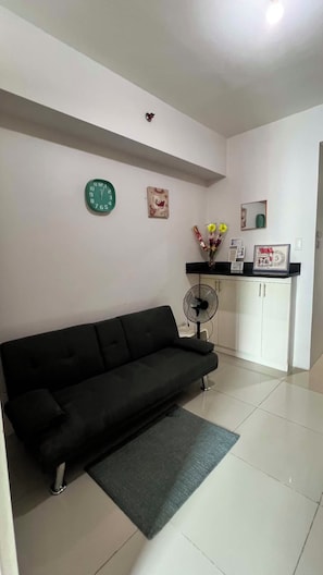 A living room with a black sofa, a small rug, a white cabinet, a fan, and a clock on the wall.