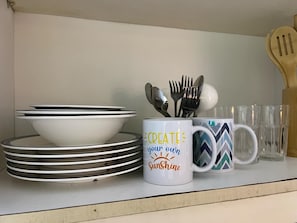 A kitchen shelf with stacked white plates, a white mug with a motivational quote, and a patterned mug. - MOA-View 1BR - 5 min Walk to MOA Arena (Pasay)