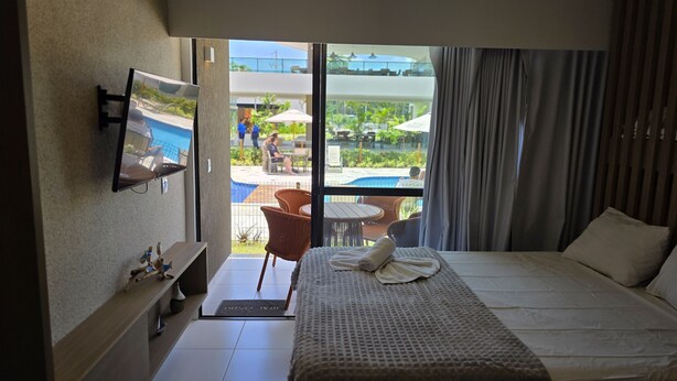 A hotel room with a bed, a TV, a small table, and a view of an outdoor pool area. - Makia Beach Exp 08A by La Playa (Ipojuca)
