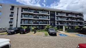A multi-story hotel with a modern design, featuring balconies on each floor, a parking area with several cars, and a sign indicating the hotel name 'KAI'. - Makia Beach Exp 08A by La Playa (Ipojuca)