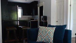 Un ambiente compatto con un divano blu scuro, un cuscino decorativo e una piccola cucina sullo sfondo.