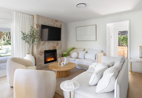 Una sala de estar moderna con chimenea de piedra, televisor de pantalla plana en la pared, sofá blanco y una mesa de centro redonda.