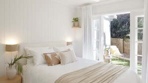 Un dormitorio con una cama grande, una mesita de noche con lámpara y vistas a un jardín a través de una puerta abierta.