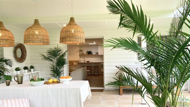 Una habitación con una mesa de comedor dispuesta, una cesta de fruta, un mantel blanco y un cojín a rayas. Se observan lámparas de mimbre colgantes, un espejo y varias plantas en macetas.