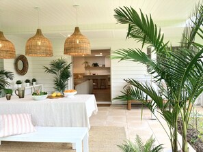 Una habitación con una mesa de comedor dispuesta, una cesta de fruta, un mantel blanco y un cojín a rayas. Se observan lámparas de mimbre colgantes, un espejo y varias plantas en macetas.
