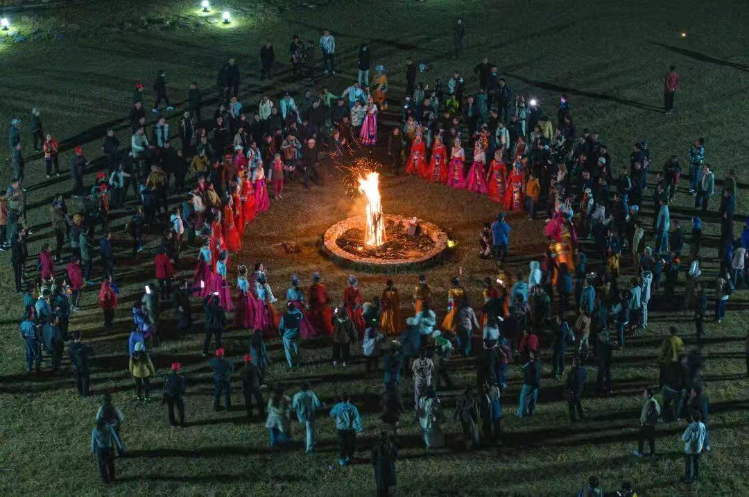 A large outdoor gathering around a central fire with people standing in a semi-circle.