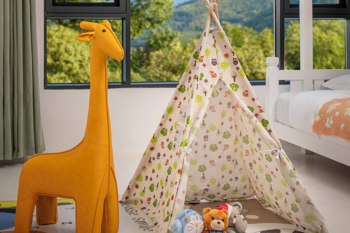 Um quarto com uma janela grande, um berço branco, um brinquedo de girafa amarela, um tipi e diversos bichos de pelúcia no chão.