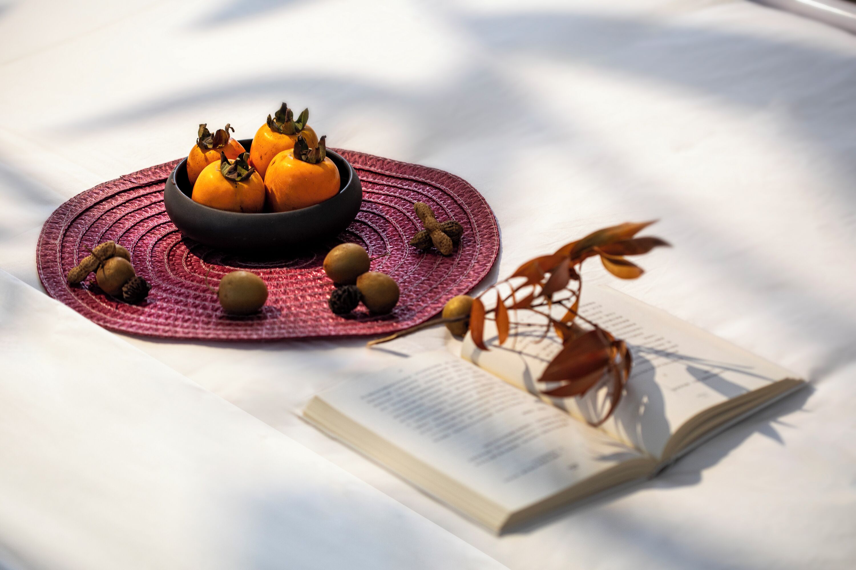 A table with a bowl of oranges, a book, and a decorative branch.