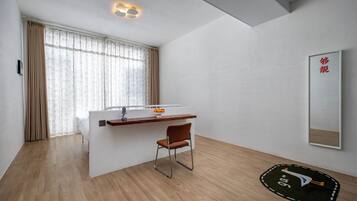 A modern hotel room with a wooden floor, a small desk with a chair, a large window with curtains, and a wall-mounted mirror.