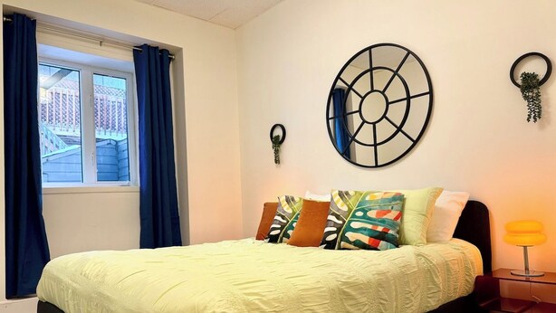 A bedroom with a bed, a window with curtains, a wall-mounted circular mirror, and a bedside table with a lamp. - Cute little apartment in the city center (Québec)