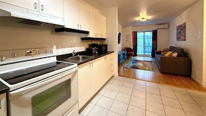 A kitchen with a stove, sink, and cabinets, leading to a living area with a couch and a sliding glass door. - Charming, well-furnished apartment (Québec)