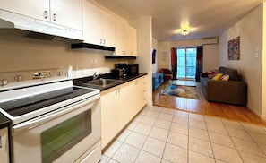 A kitchen with a stove, sink, and cabinets, leading to a living area with a couch and a sliding glass door.