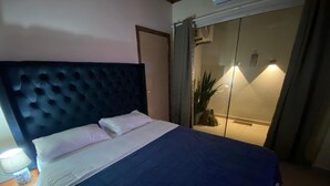 A bedroom with a large bed, a nightstand, a lamp, and a sliding glass door leading to a balcony with a plant.