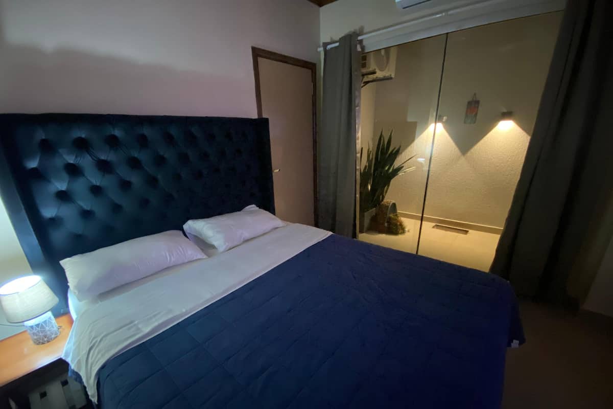 A bedroom with a large bed, a nightstand, a lamp, and a sliding glass door leading to a balcony with a plant.