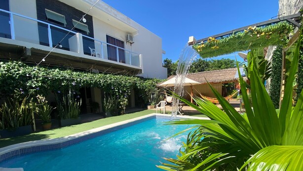 A swimming pool with a water feature, surrounded by lush greenery and a building with a balcony. - Hotel Junior (Caazapá)