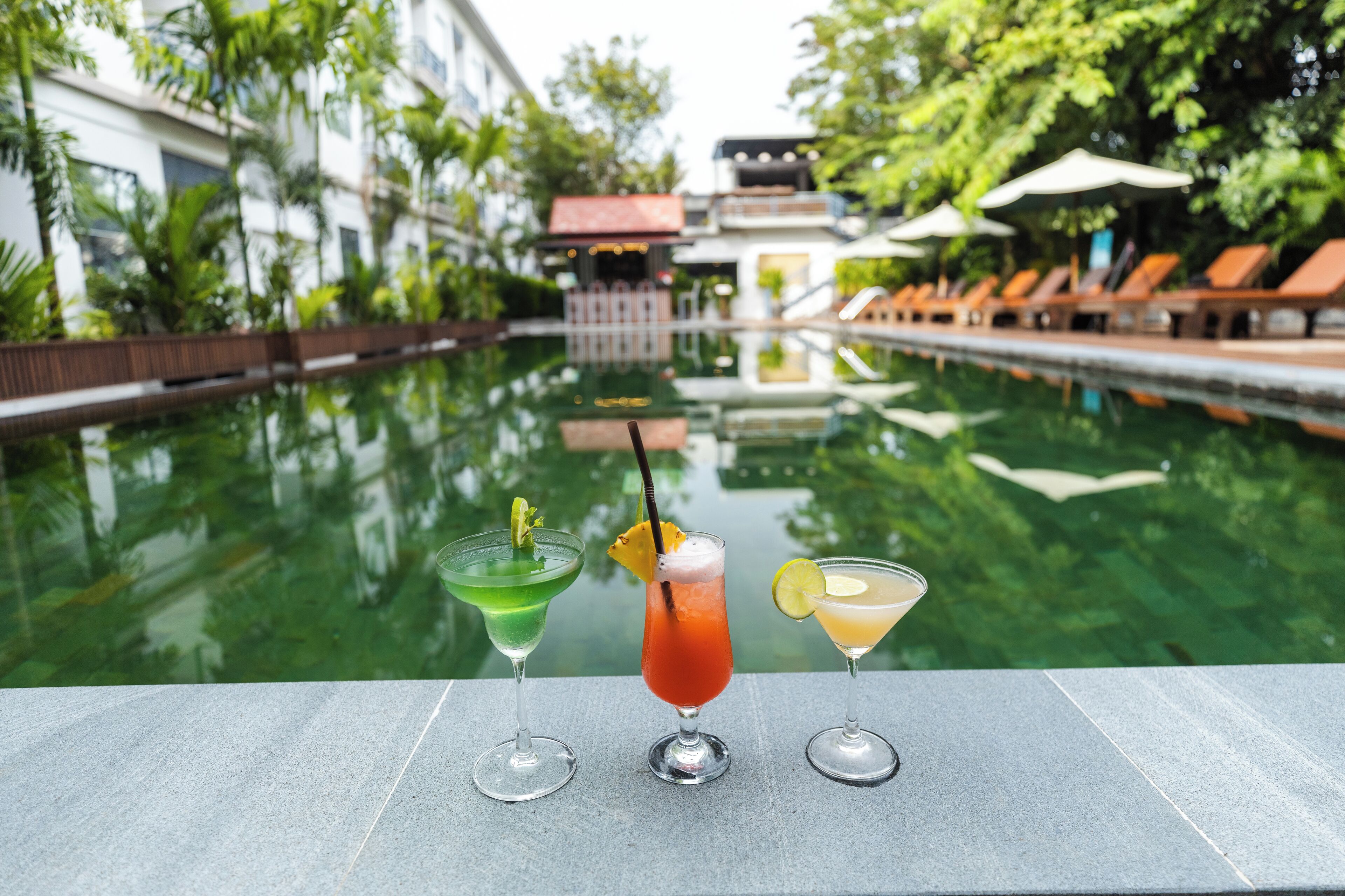 Un espace aménagé au bord de la piscine, avec trois cocktails, le reflet d’un bâtiment et des chaises longues.