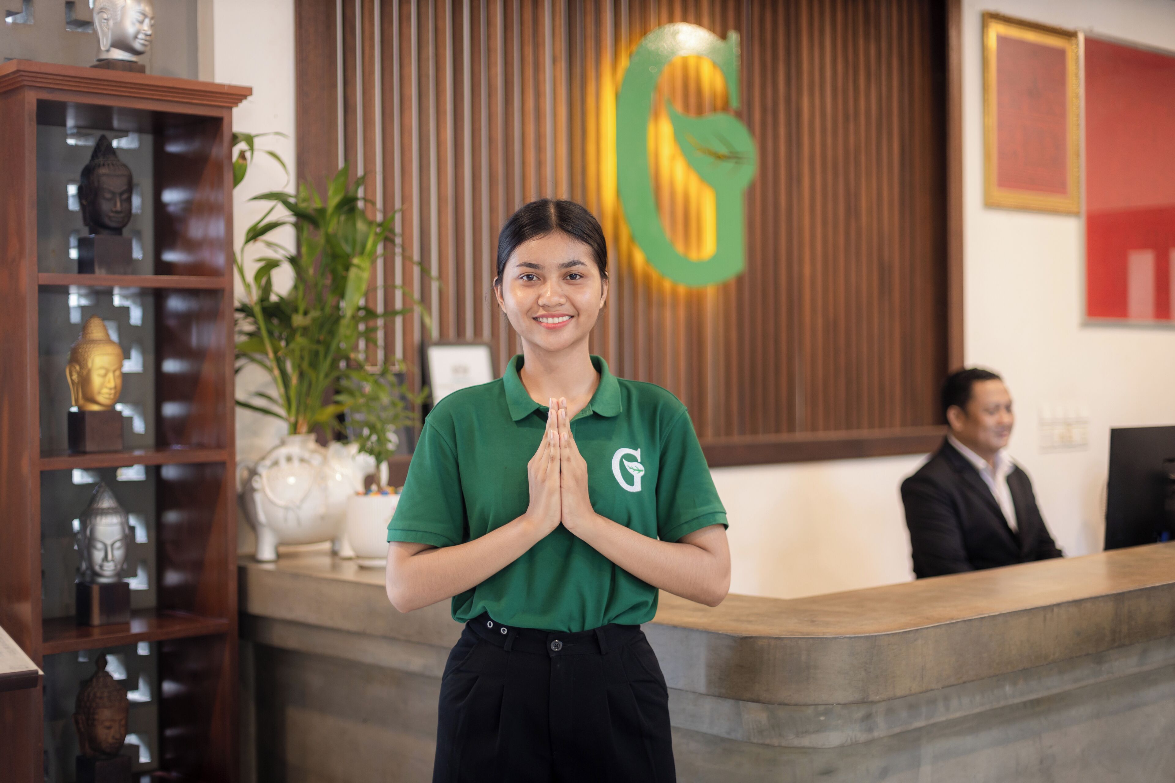 A reception area with a wooden reception desk, a Buddha statue, and a green logo on the wall.