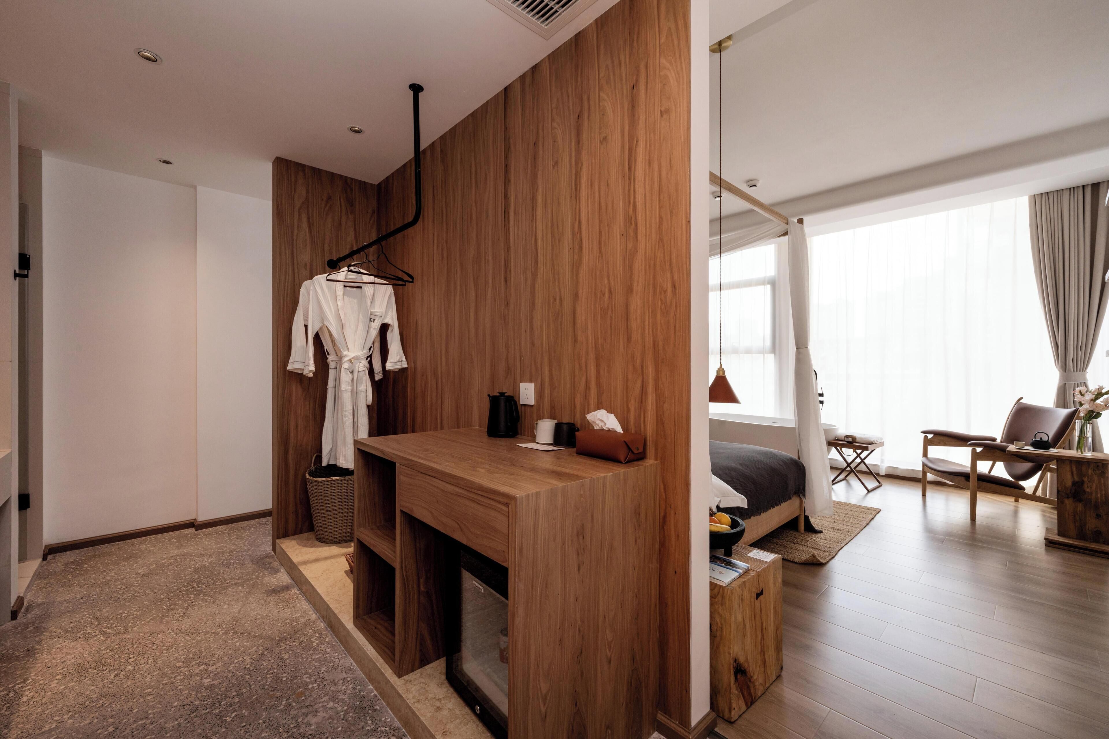 A modern hotel room with a wooden partition, a small desk with a kettle, a hanging robe, and a bed with a view of the city.