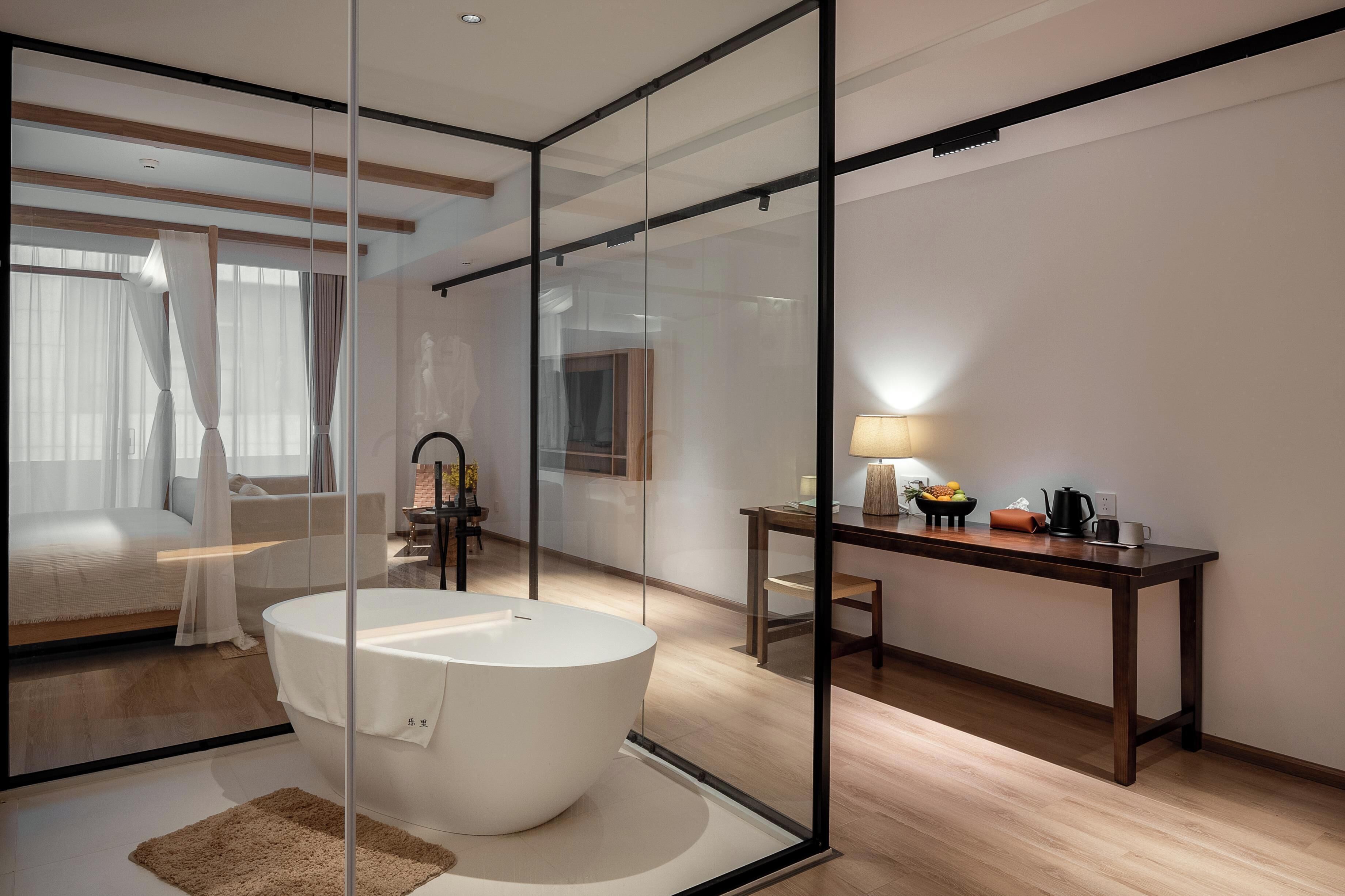 A modern hotel room with a large bathtub, a wooden desk, and a view through a glass partition.