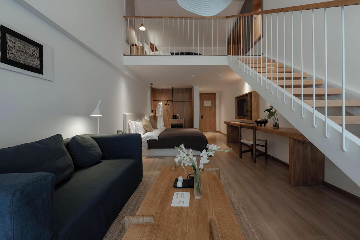 Een moderne woonkamer met een donkerblauwe bank, een houten salontafel met een bloevas, een trap met een witte leuning, en een eethoek van hout.