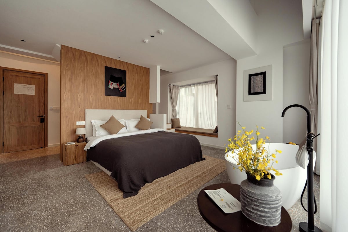 Een moderne hotelkamer met een groot bed, een nachtkastje met een lamp, een raam met gordijnen, een aan de muur hangend schilderij en een bloemenvaas op een bijzettafeltje.