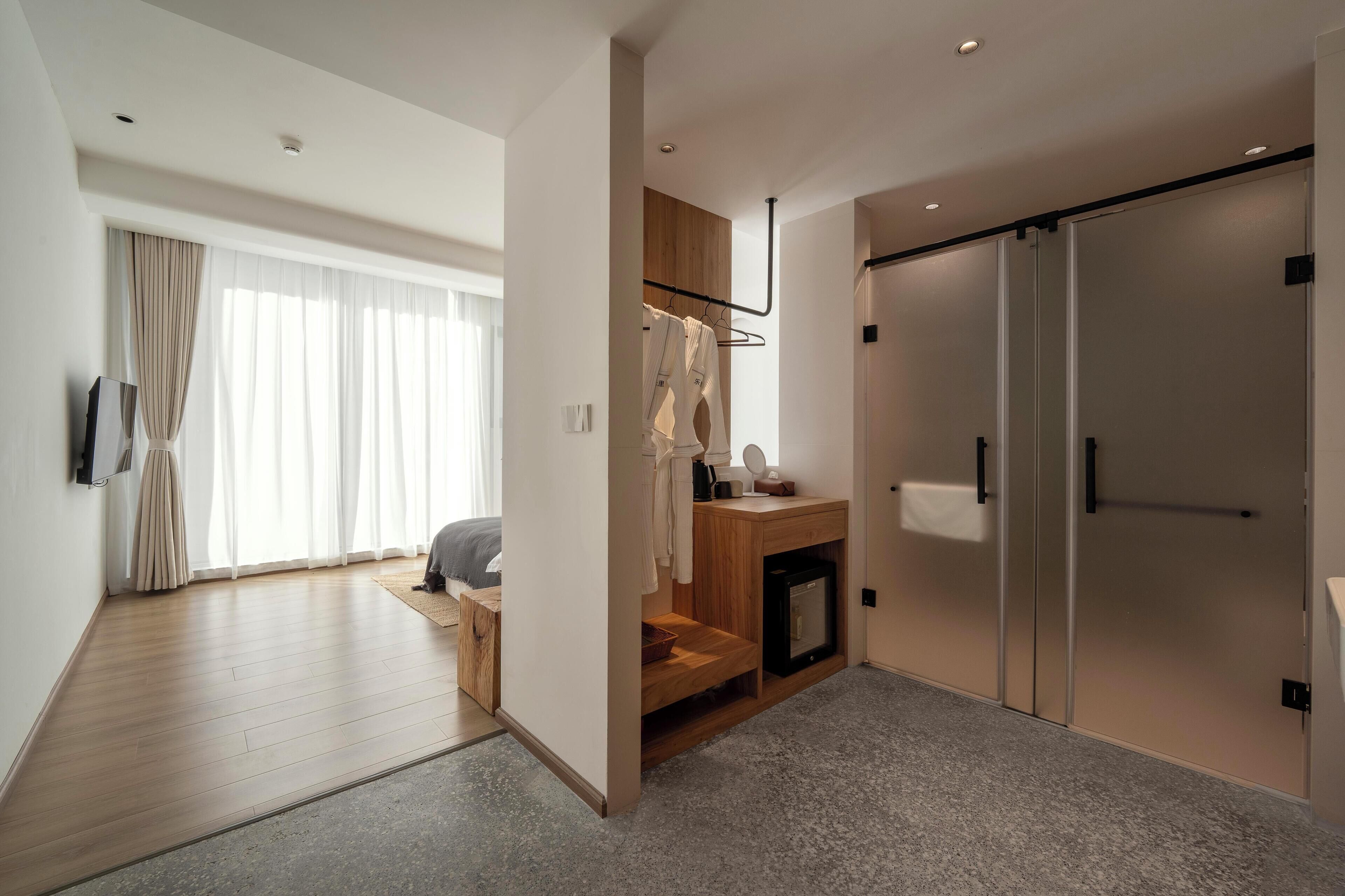 A modern hotel room with a wooden floor, a small seating area, a built-in wardrobe, and a large window with sheer curtains.