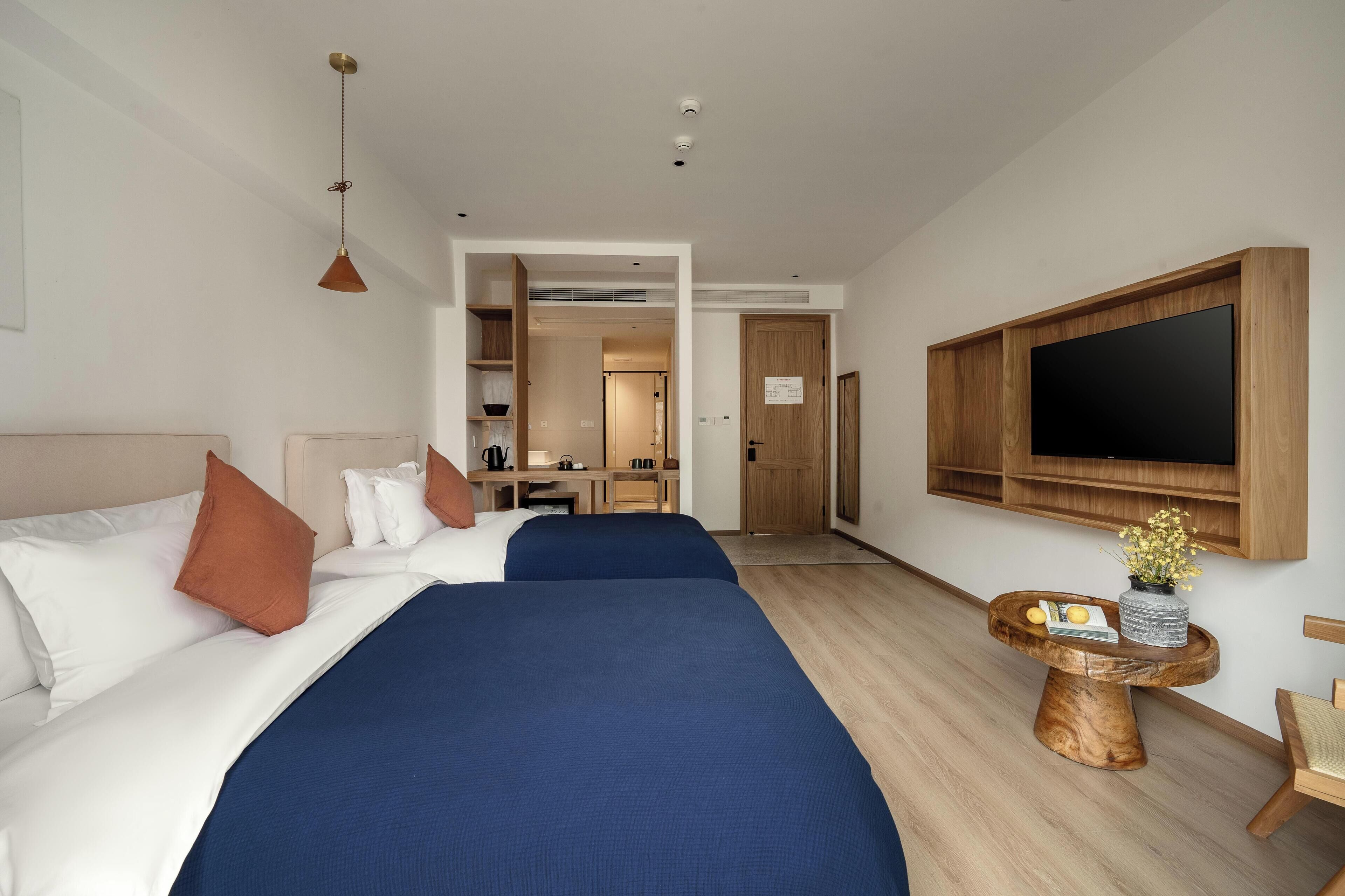A hotel room with two beds, a TV mounted on the wall, a small wooden side table with a vase, and a kitchenette in the background.