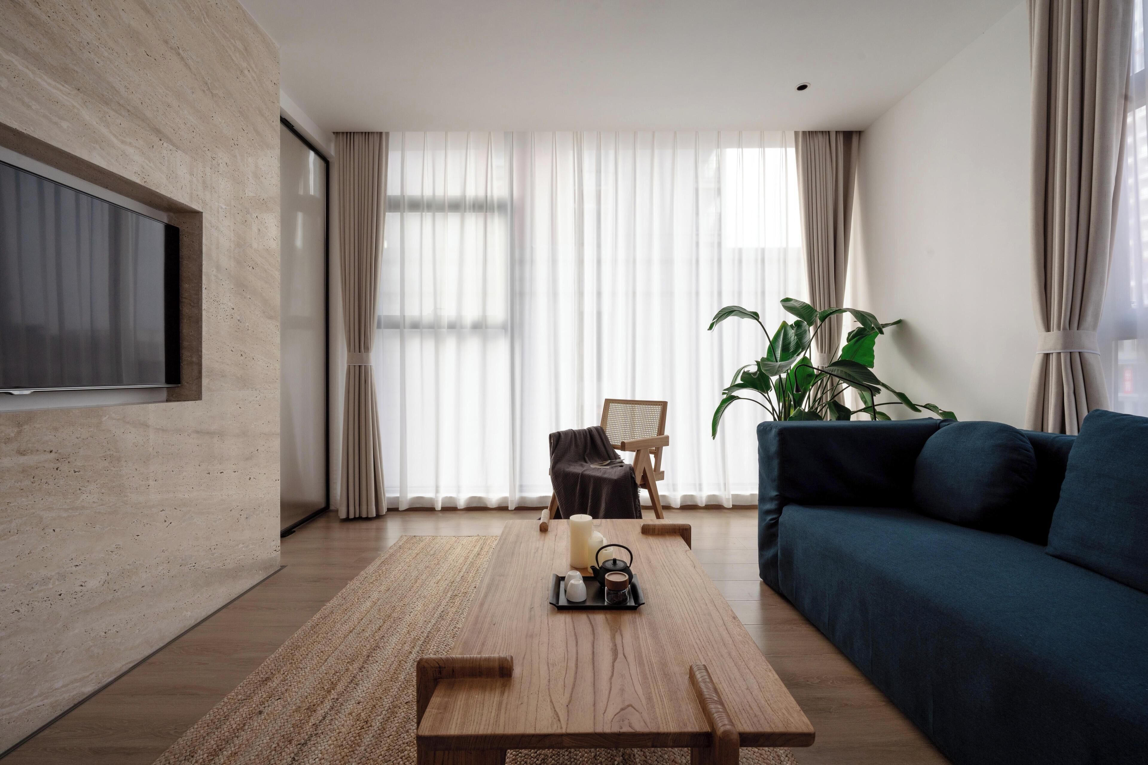 A modern living room with a wooden coffee table, a blue sofa, a TV mounted on a textured wall, and a large window with sheer curtains.