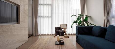 A modern living room with a wooden coffee table, a blue sofa, a TV mounted on a textured wall, and a large window with sheer curtains.