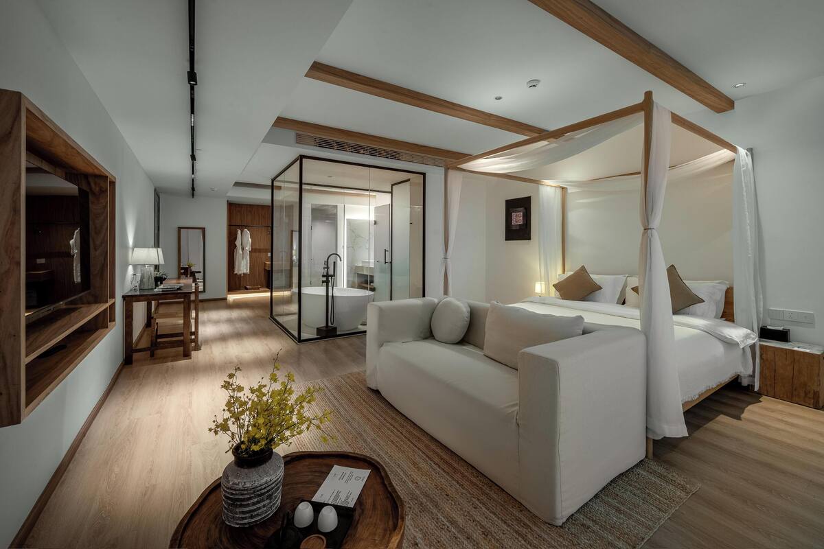 Een moderne hotelkamer met een hemelbed, een bank, een bureau en een badkamer met glazen wanden.