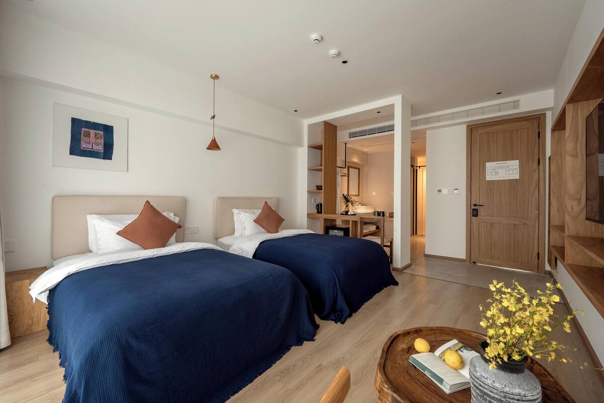 Een hotelkamer met twee bedden, een houten vloer, een bureau met stoel en een tafeltje met een bloevas.