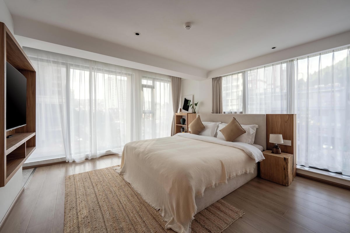 Een moderne slaapkamer met een groot bed, een houten hoofdbord, nachtkastjes, een televisie en dunne gordijnen.