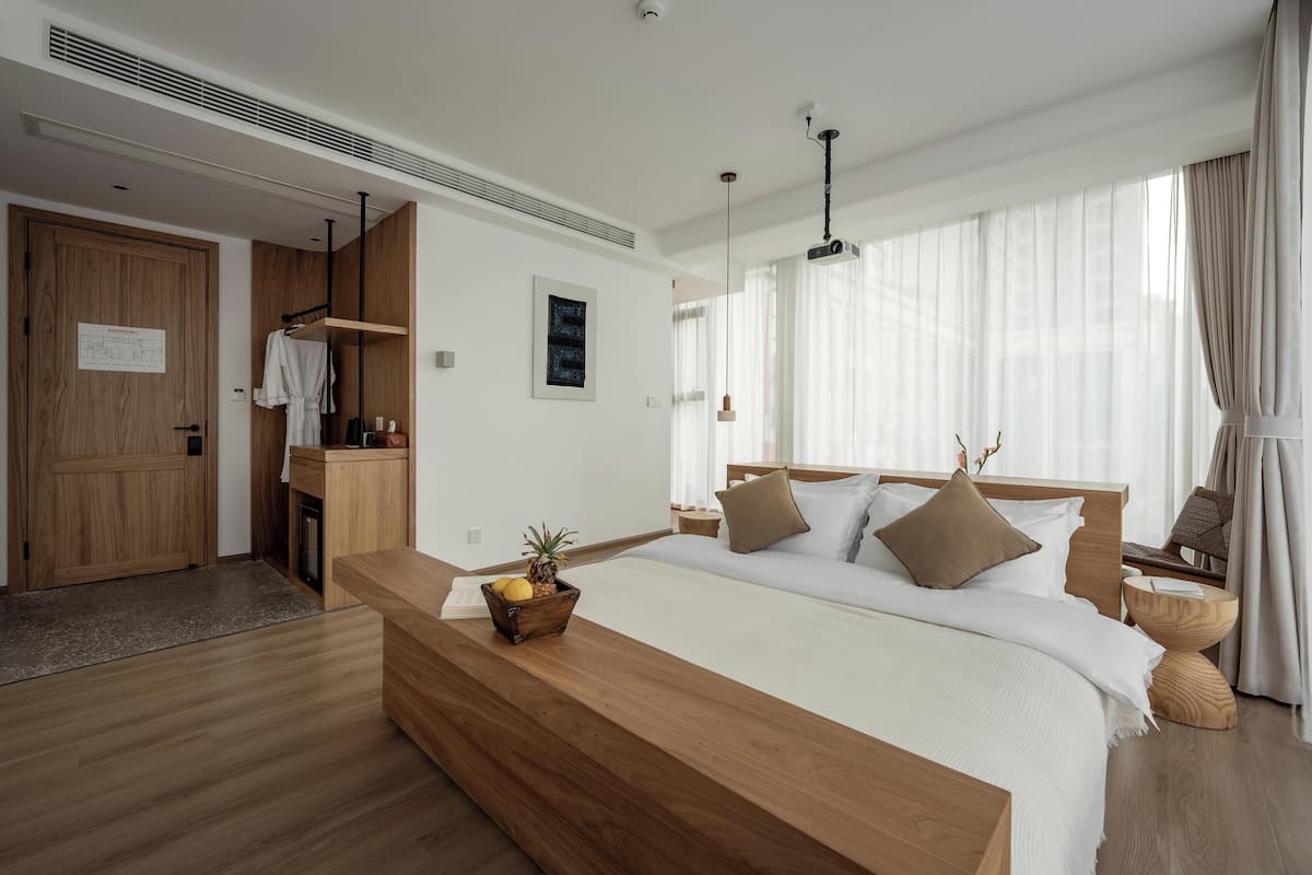 Een moderne hotelkamer met een houten bed, een houten bankje met een fruitmand, een klein houten tafeltje, een stoel en een groot raam met dunne gordijnen.