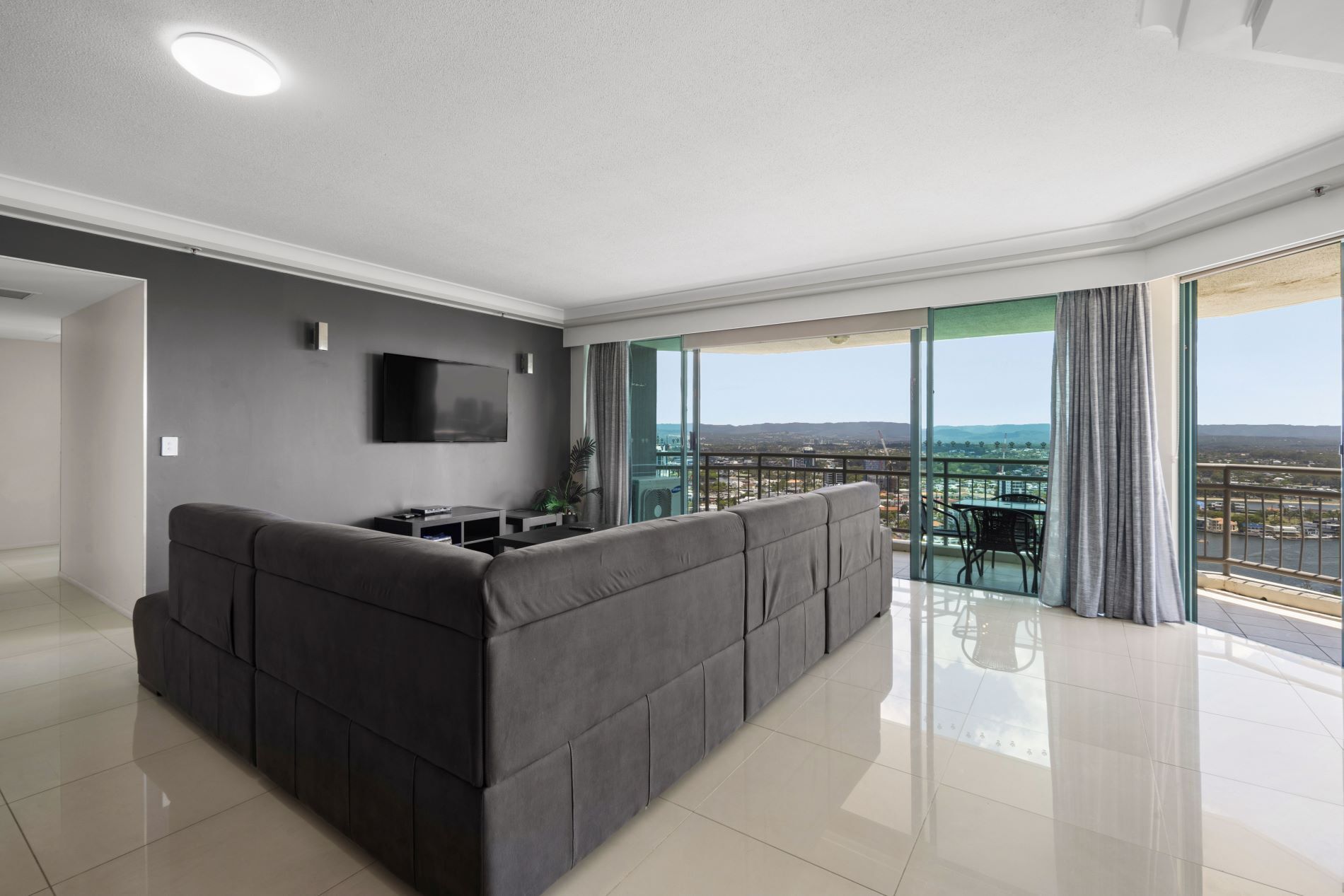 Una sala de estar moderna con un sofá grande de color gris, un televisor de pantalla plana montado en la pared y un balcón con vistas a la ciudad.