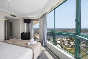 A hotel room with a large window offering a city view, a bed with a beige blanket, a black cabinet, and a flat-screen TV. - Crown Towers Resort - Luxury Apartment (Surfers Paradise)