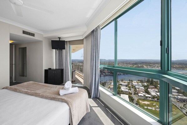 Habitación de hotel con un ventanal grande que ofrece vistas a la ciudad, una cama con manta color beige, un mueble negro y un televisor de pantalla plana.
