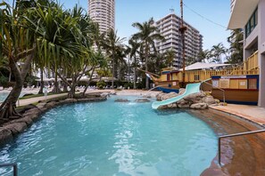A pool area with a water slide, palm trees, and a building in the background. - Crown Towers Resort - Luxury Apartment (Surfers Paradise)
