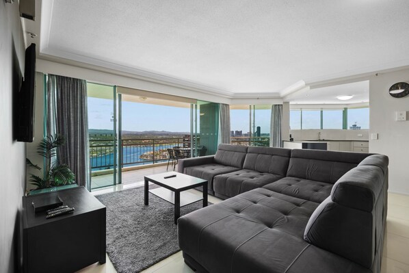 A modern living room with a grey sofa, a small table, and a TV. There is a balcony with a view of a body of water and buildings.