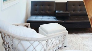 Una sala moderna con un sofá de cuero negro, una silla de mimbre blanca y un reposapiés blanco. Se observa una ventana con persianas, un ventilador de techo y un equipo de aire acondicionado.