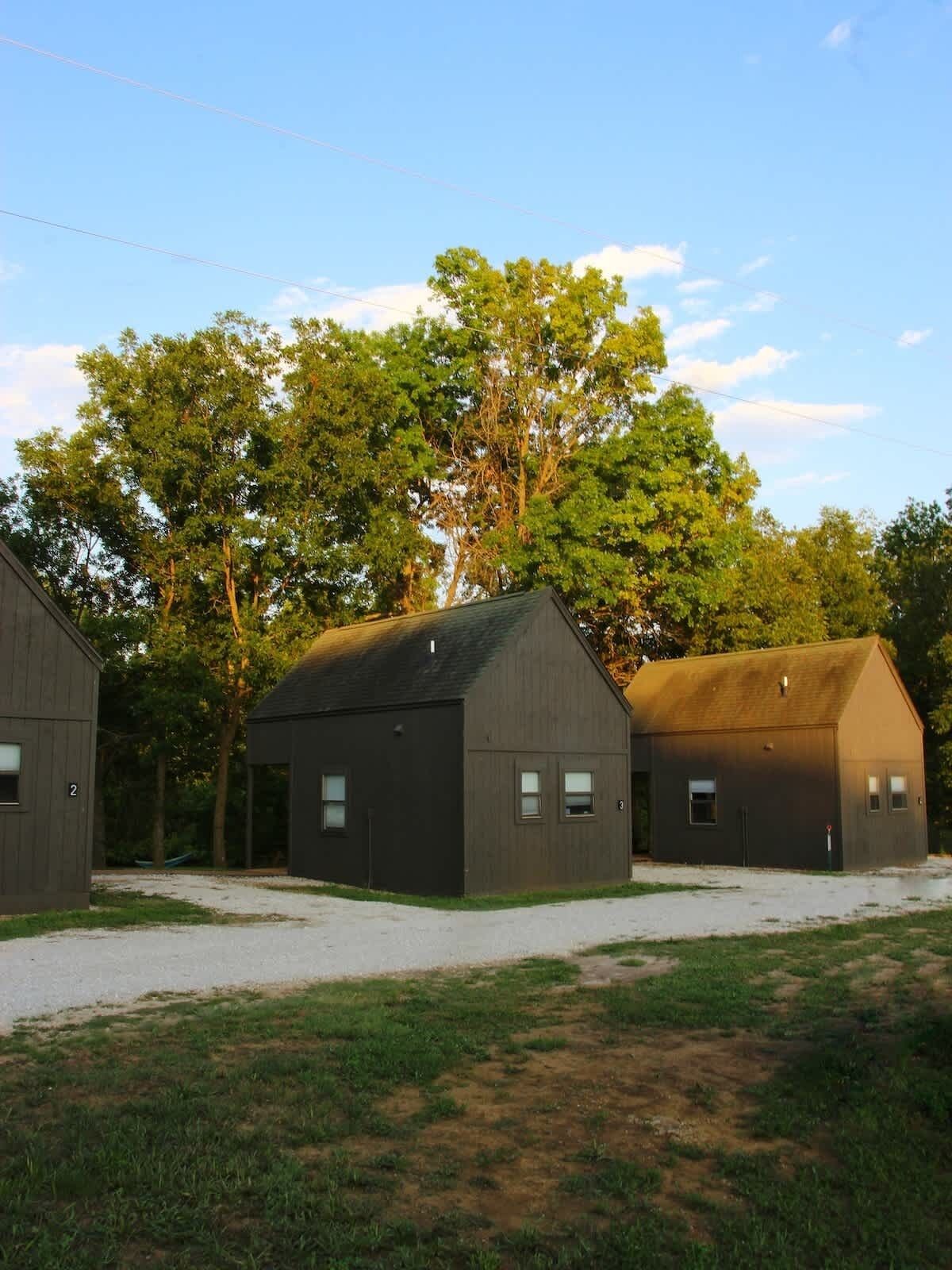 Drei Holzgebäude mit nummerierten Fenstern, vor einer Baumkulisse und blauem Himmel.