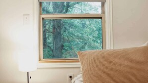 Quarto com cama, mesa de cabeceira com abajur, janela com persianas e vista para árvores.